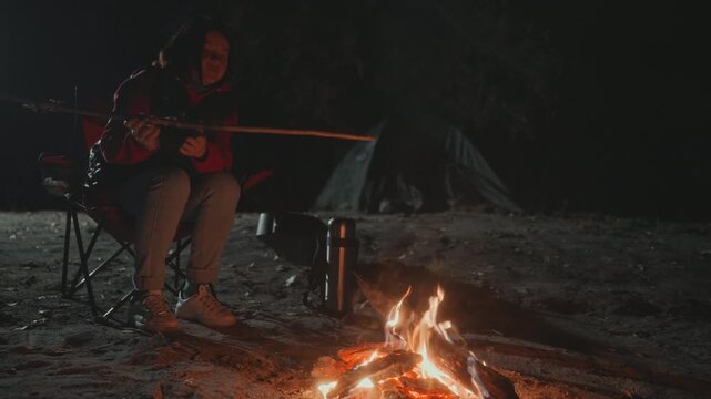 Solo camper cooking over campfire at night folding chair and tent backdrop, long stick used to roast snack, metal kettle and pot near flame, person in red jacket tending fire, sparks rising into dark