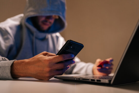 Close-up of hooded person engaging in cybercrime, performing online fraud and data theft using mobile phone, laptop and credit card in dark environment, representing internet security threats.