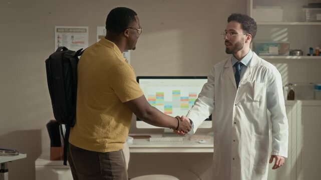 Medium shot of Middle Eastern male doctor in white lab coat and glasses greeting Black male patient during appointment in modern clinic office with desktop computer
