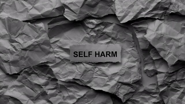A torn piece of paper with the words Self harm on it lying on a background made of chaotic crumpled paper textures. Black and white. Close up.