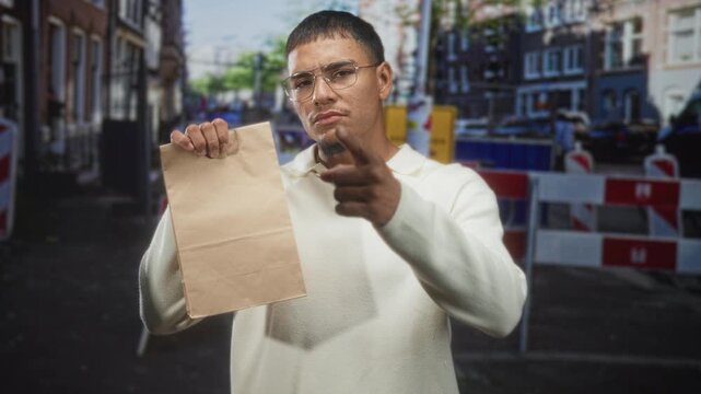 Man holds paper bag in one hand and points finger toward camera on a street, wearing glasses and a white sweater; assertive demand.