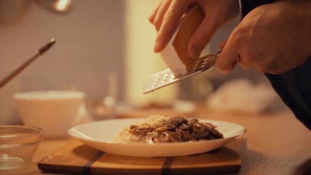 A man, acting as the kitchen's chef, grates hard Parmesan with a hand grater onto a steaming plate of cooked food, filling the evening cooking space with aromatic cheese and culinary focus