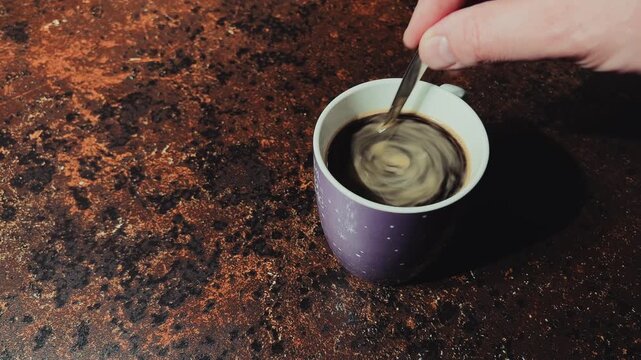 A hand stirs black instant coffee with a metal spoon in a purple ceramic mug. Close-up shot on a dark brown textured surface in natural light.