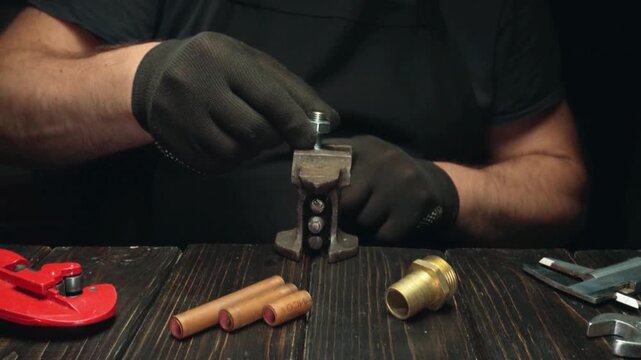Male technician in black gloves tightens a metal bolt using a tool on a workbench with copper pipes and various tools arranged nearby