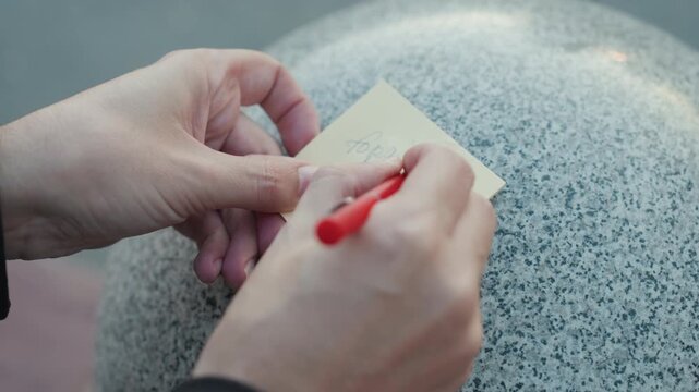 Closeup hands writing note on granite, red pen on sticky paper against rounded bollard coat sleeve visible, deliberate strokes, quick doodle and listmaking, planner leaving reminder, quiet urban