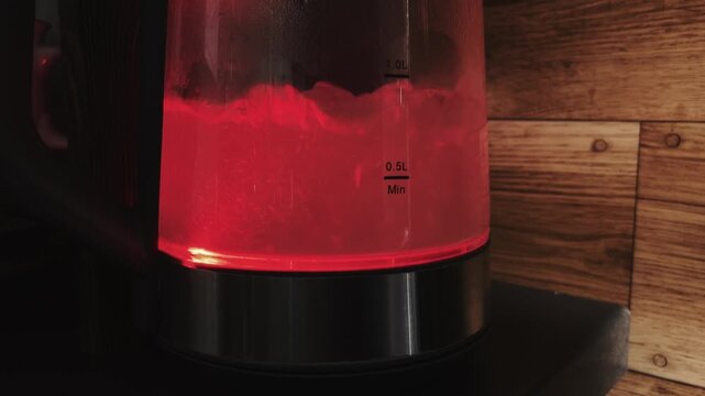 Static shot of a glass electric kettle with boiling water inside. The water is illuminated by a bright red LED light at the base. Wooden wall background.