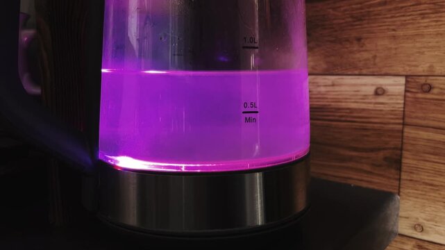 Close-up of a glass electric kettle with purple LED illumination heating water. Boiling process visible through transparent walls against a wooden background.
