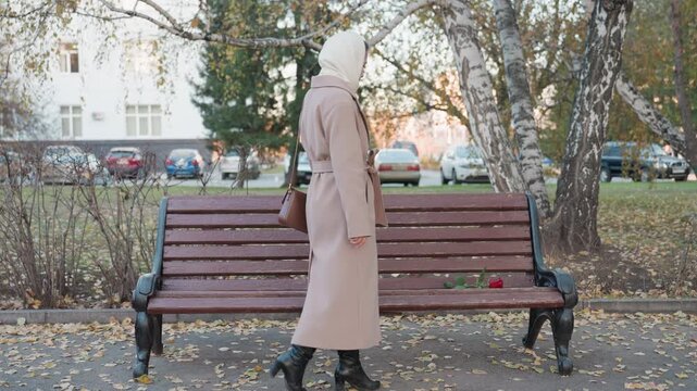 Caucasian woman places single red rose on wooden bench in autumn park, steps back and pauses while wrapped in pale coat and headscarf wide framing reveals birch trees, parked cars, scattered leaves