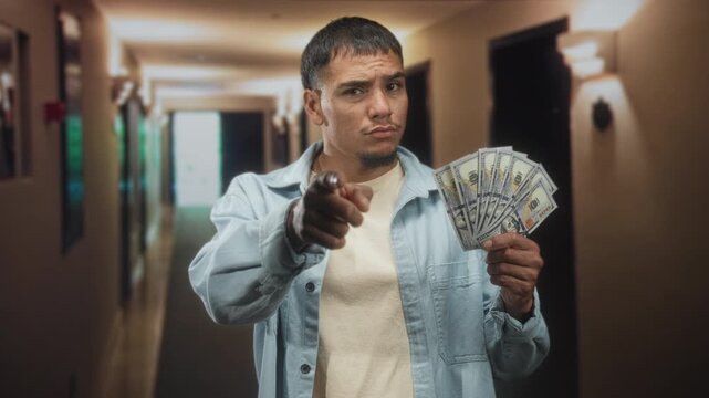 Young hispanic man holding us dollar bills and pointing finger in a hotel corridor with wall sconces and doors visible; bravado confidence.
