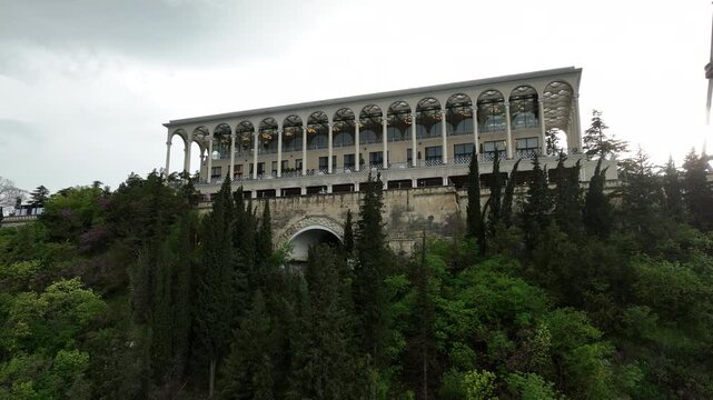 Aerial view of Mtatsminda Park funicular station and restaurant building in Tbilisi, Georgia, with the TV tower and Ferris wheel in a lush green mountain landscape.