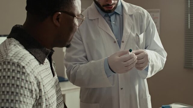 Medium shot of Middle Eastern male healthcare professional holding insulin pen while explaining diabetes management to Black male patient in clinic office