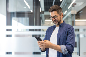 Fototapeta premium Smiling man with glasses using smartphone in a modern office, networking and texting during the workday, confident professional connecting via mobile apps and social media, waist-up portrait