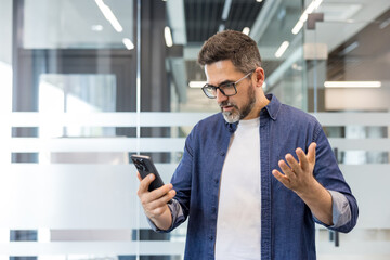 Fototapeta premium Mature professional holding smartphone, looking frustrated and confused after reading bad news or an upsetting message in a modern office environment, stressed and pensive