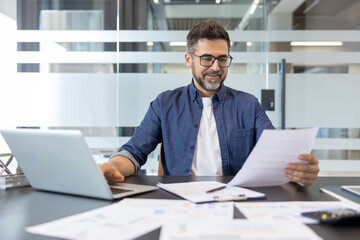 Obraz premium Professional man wearing glasses and a denim shirt sitting at his office desk, happily engaged in work while using a laptop and reviewing financial documents with graphs on the table
