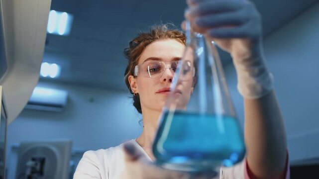 Laboratory technician studying blue solution in flask under laboratory lighting. Concept biomedical science, infectious disease research and virology including COVID-19 coronavirus and hantavirus.
