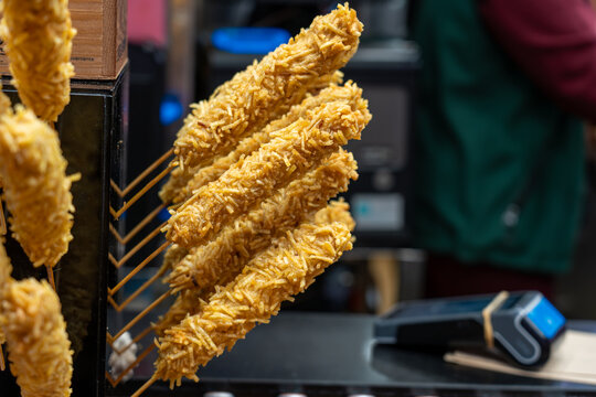 Close-up of vertical skewers of deep-fried snacks wrapped in crispy potato shreds, displayed at a food market.