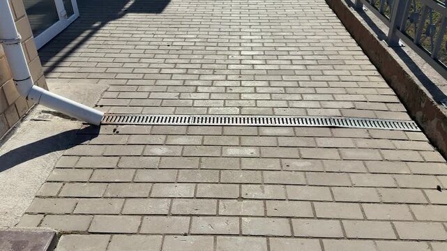 White downpipe and metal grate on paving stones, sunlight casting shadow across textured pavers, closeup view of storm water
