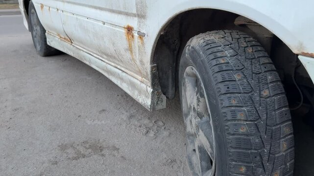 White car side view with rust, punctured front wheel and muddy studded tire parked on roadside pavement visible corrosion