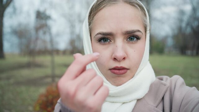 Young woman in scarf sternly beckoning, pointed finger and intense stare in winter park, closeup selfie capturing confrontational energy, bare trees and coat emphasize cool mood, authentic social