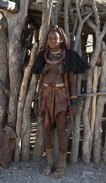 Young traditionally dressed Himba woman, leaning against a wooden hut, near Opuwo, Kaokoveld, Kunene, Namibia