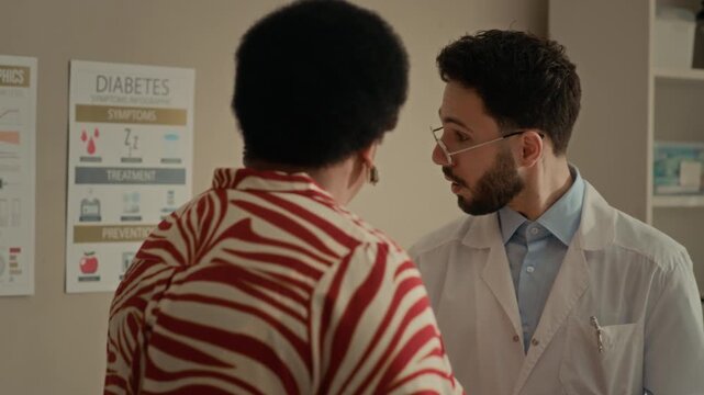 Back view shot of physician explaining diabetes symptoms and treatment information to senior Black female patient using educational posters in medical office