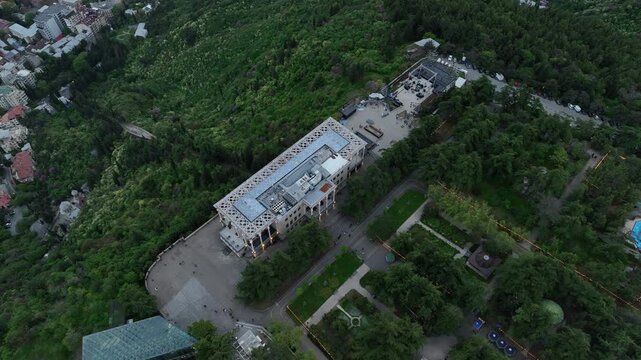 Aerial view of Mtatsminda Park funicular station and restaurant building in Tbilisi, Georgia, with the TV tower and Ferris wheel in a lush green mountain landscape.