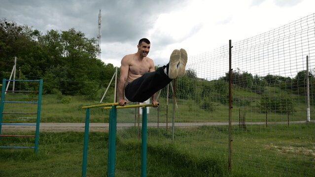 Strong man trains abdominal muscles raising legs on parallel bars at sports ground. Young shirtless sportsman exercising at nature. Sporty athlete working out at nature. Concept of sportive lifestyle