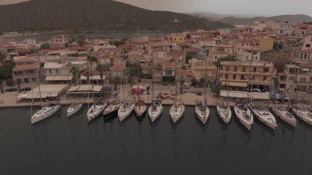 Smooth aerial drone flyby from left to right parallel to docked sailing yachts in Ermioni harbor under dramatic Mediterranean orange sky