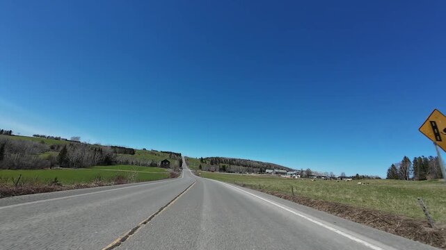 4K dashcam footage of a vehicle speeding along a rural road through farmland and passing an abandoned old house beneath a clear blue sky in Gasp&eacute; Peninsula, Quebec, Canada.