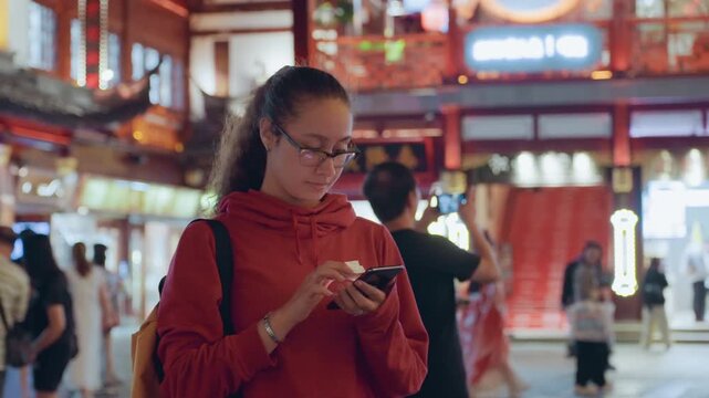 Young woman in red hoodie texting in temple square, checking map app, navigating crowds, observing architecture, evening neon glow, thoughtful traveler moment, backpack and glasses visible, urban calm