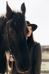 Fototapeta premium Woman stands next to a black horse in a rural area during the day while holding the reins and wearing a hat