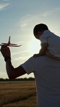 White Father With Toy Airplane On Shoulders At Golden Sunset Field. Father Plays Pilot With Toddler On Shoulder Ride, Hand Guiding Red Model Airplane Into Backlit Sky, Warm Glow, Gentle Breeze, Quiet