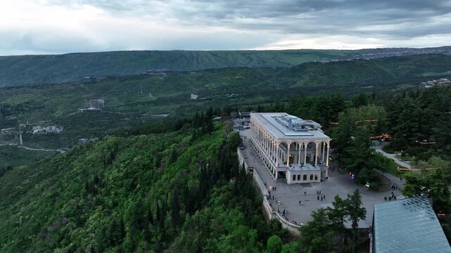 Aerial view of Mtatsminda Park funicular station and restaurant building in Tbilisi, Georgia, with the TV tower and Ferris wheel in a lush green mountain landscape.