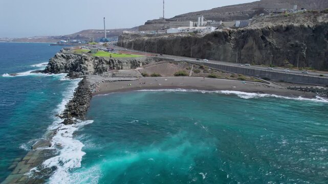 Beautiful aerial perspective of the volcanic La Laja beach in Gran Canaria. Turquoise ocean waves crashing against the rocky shore and breakwater