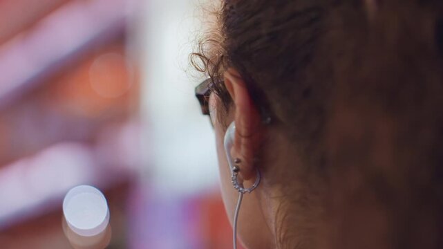 Close profile adjusting earring under neon lights, hair in a bun and patterned glasses, small hoop piercings and attentive gesture, candid urban night scene with colorful bokeh and intimate detail.
