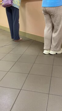 Two women at reception desk rear view, standing side by side on tiled floor, casual clothing and tote bag, muted pastel counter