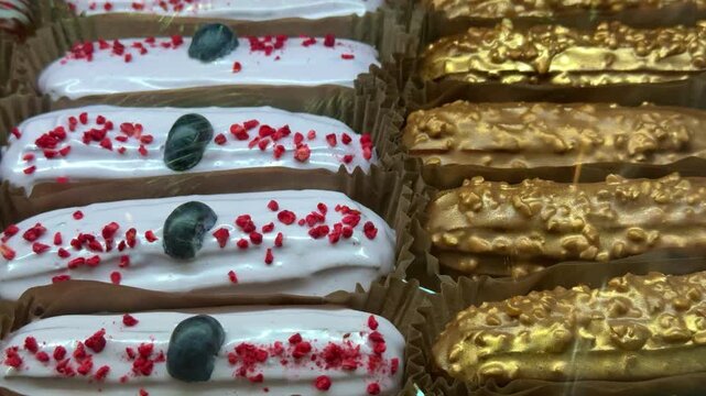 Close-up display of eclairs arranged in coffee shop glass case, white iced eclairs topped with red sprinkles and dark berry