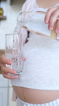 Close up of a pregnant woman pouring pure, fresh water from a glass pitcher into a tall glass, with her baby bump and linea nigra visible, showcasing the importance of hydration during pregnancy