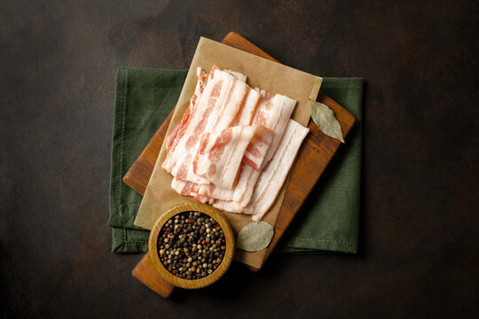 Raw bacon on wooden cutting board on brown background. Streaky brisket slices, fresh thin sliced bacon. Top view