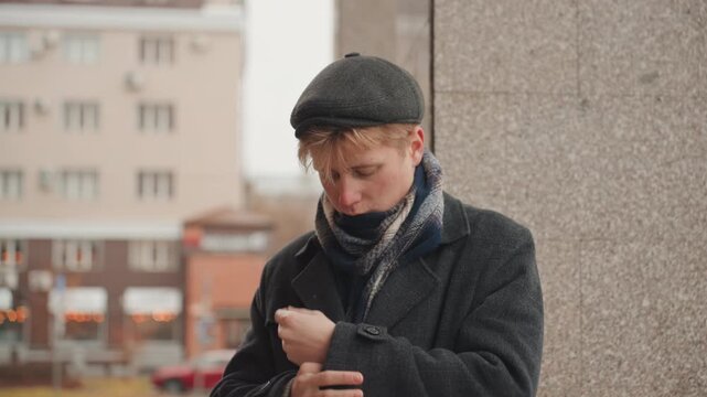 Young white creator adjusts scarf coat outdoors hands fumble with collar against concrete column, muted city buildings in background, vintage cap and layered outerwear convey chilly mood and creative