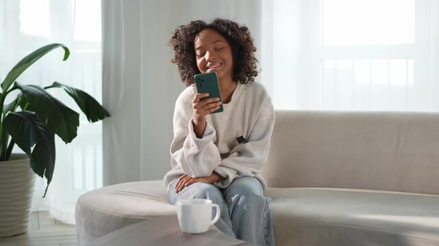African girl holding smartphone touch screen typing scroll page at home. Woman with cell phone surfing internet using social media apps playing game. Shopping online Internet news cellphone addiction