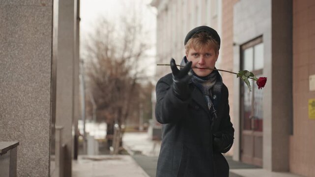 Young white creator playful with rose on chilly city sidewalk, wearing coat, scarf, beanie and leather gloves, teasing smile, miming kiss, cheeky gestures toward camera, cinematic candid portrait
