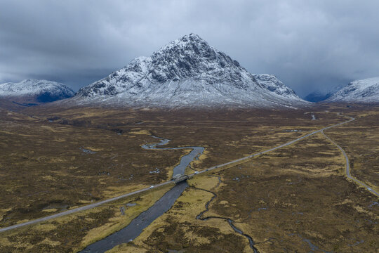 Aerial view of River Etive Bridge crossing the winding river with the snow-capped Buachaille Etive Mor mountain under a moody sky Ballachulish, Scotland, United Kingdom.
