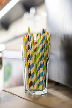 Colorful striped paper straws in a clear glass. Glass sits on a wooden surface. Background shows blurred kitchen appliances. Soft lighting highlights straws' vibrant pattern