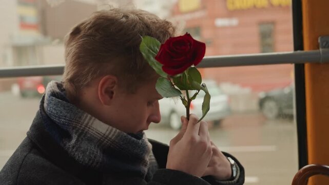 Young white creator holding red rose, on tram window seat, scarf bundled, city street passing in rain, pensive profile gaze, hands clutching stem, cinematic winter light, romantic solitude, creator