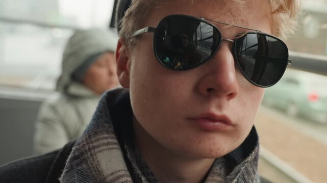 Young white creator side profile on bus window, sunglasses and beanie, layered scarf and muted city view traveling musician vibe, quiet planning, cinematic texture and subdued color