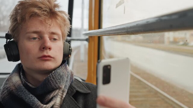 Creator man on tram checking phone, wearing overear headphones, neutral expression while scrolling social feed, raindrops on window, urban tracks passing by, candid mobile portrait, subtle motion,