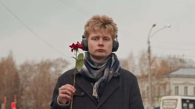 White man headphones with red rose, street backdrop and wool scarf, playful creator grin and middle finger gesture, wry humor and confident charm, candid urban portrait with warm undertone and content
