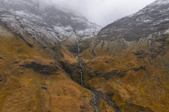 Aerial view of Glencoe valley with a thin waterfall cascading down rugged brown slopes under snow-dusted peaks and misty clouds Glencoe, Scotland, United Kingdom.