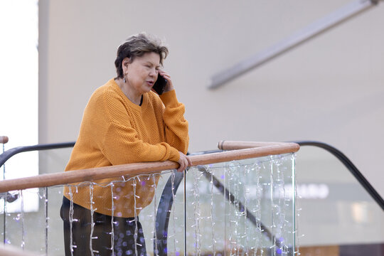 Mature woman in yellow sweater speaking on mobile phone, gesturing with hand, emotional conversation in modern interior.